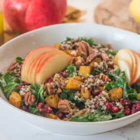 a kale salad in a white salad bowl, topped with apples, squash, and pecans