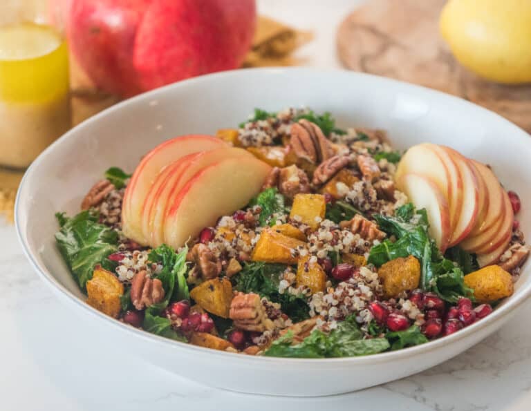 a kale salad in a white salad bowl, topped with apples, squash, and pecans