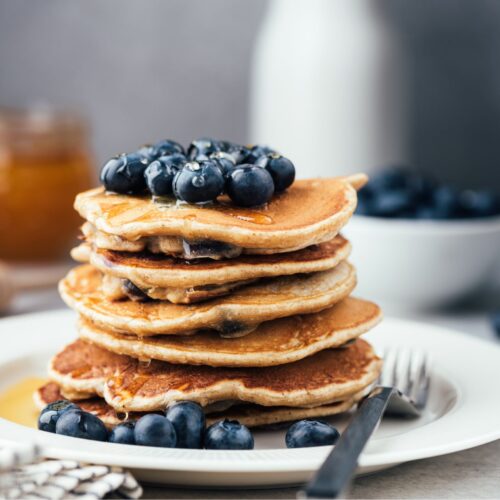 stack of blueberry pancakes sitting on a white plate, topped with blueberries