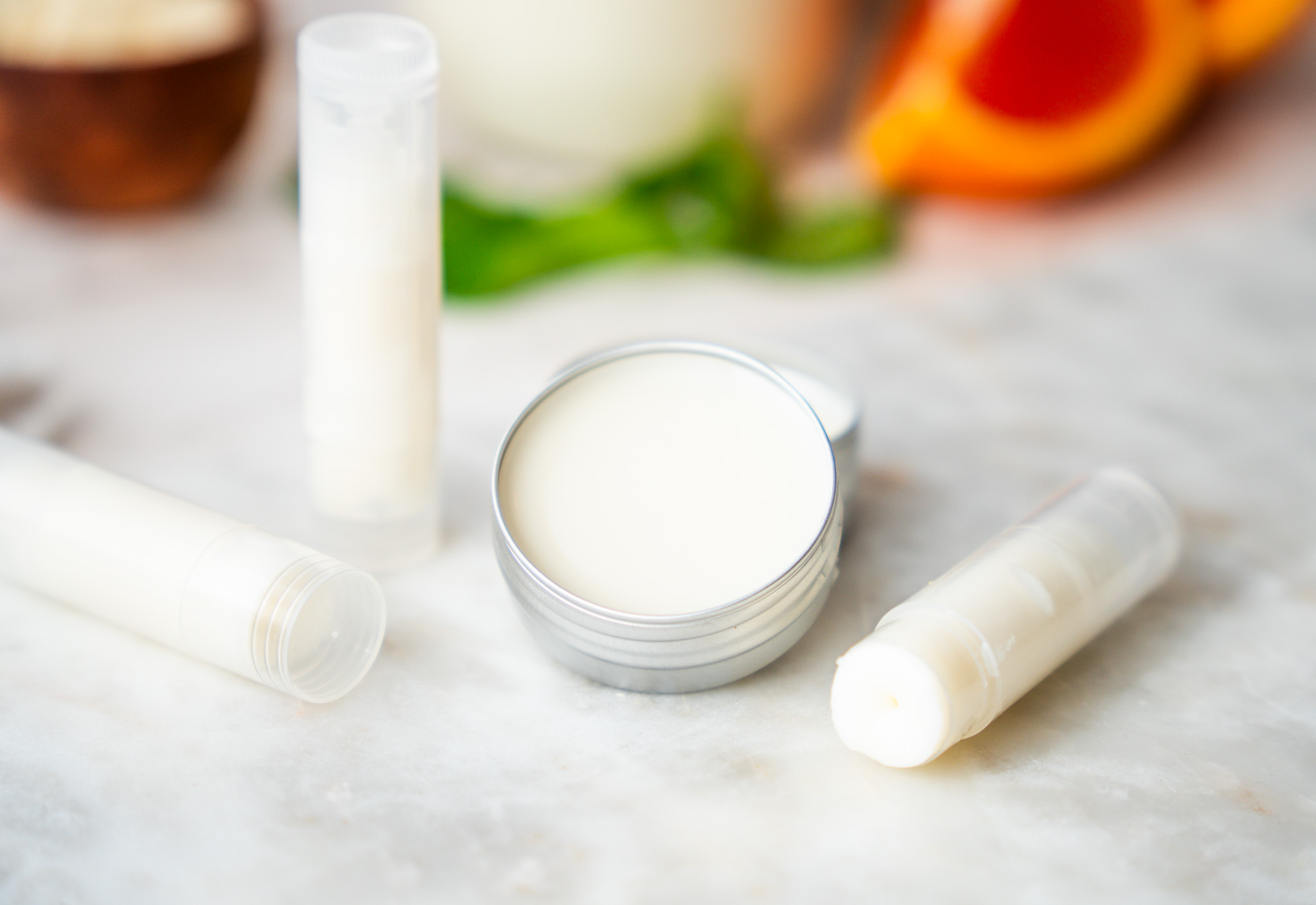Two white lip balm tubes and an open metal tin of pale homemade balm on a light marble surface with greenery behind